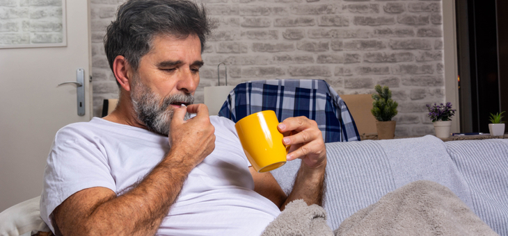 man on couch under blanket taking pill with drink