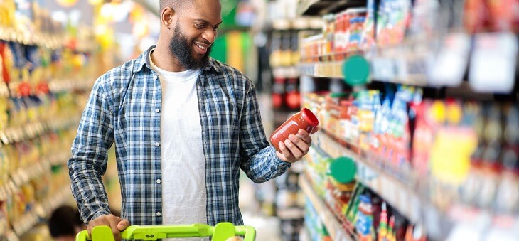 man looking at jar of pasta sauce while pushing trolley in supermarket aisle
