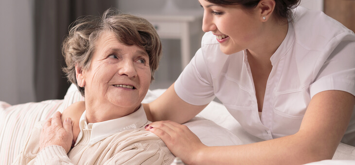 smiling nurse with hands on shoulder of smiling older woman sitting on couch