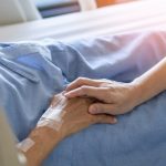 hand of palliative care nurse holding hand of older patient in bed
