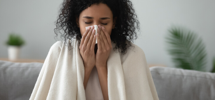 sick woman wrapped in blanket on couch blowing nose