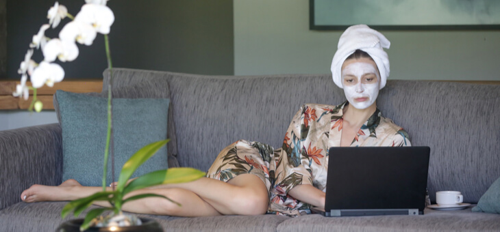 woman wearing face cream lying on couch looking at laptop