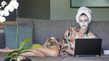 woman wearing face cream lying on couch looking at laptop