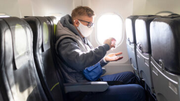 man in face mask sitting on plane applying hand sanitizer