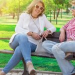 two women sitting on park bench wearing jeans