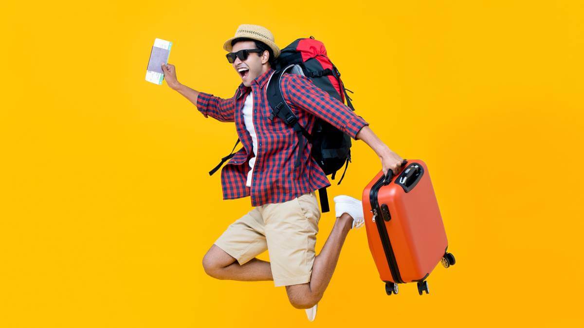 Tourist jumping in the air holding a suitcase and backpack on yellow background