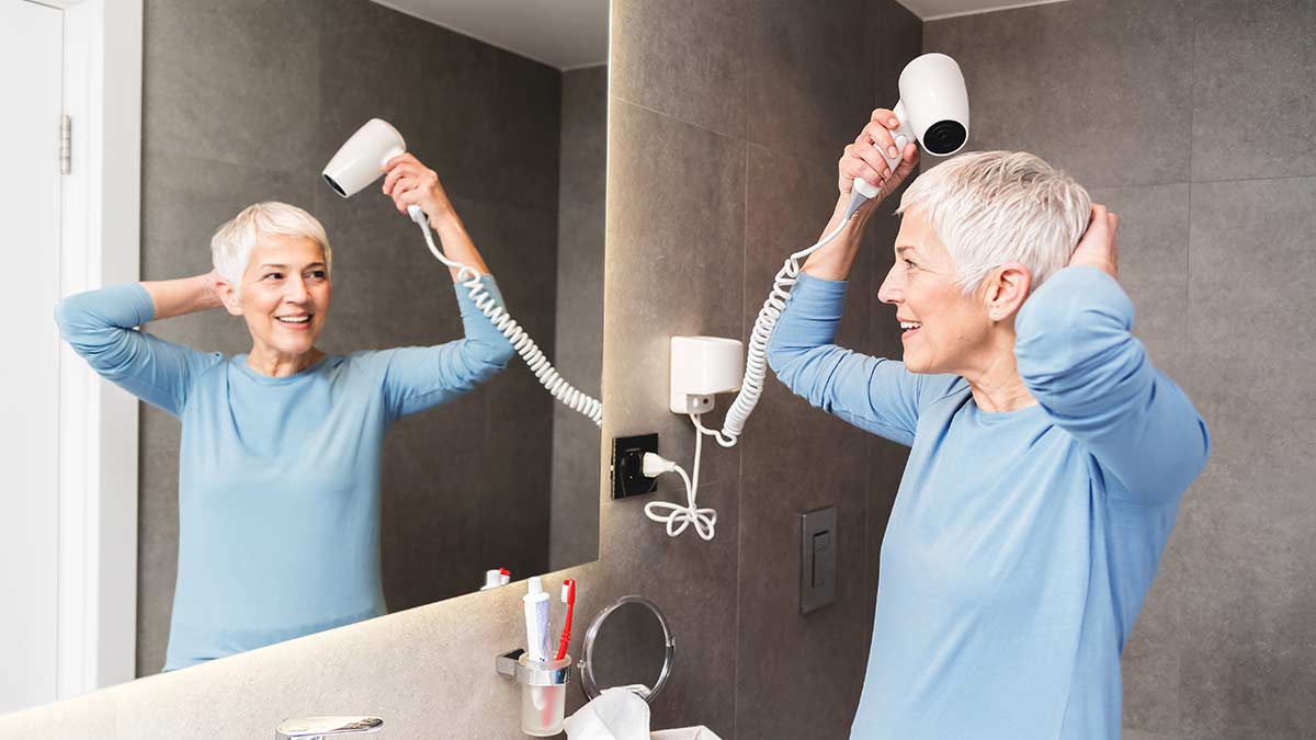 mature woman drying her short grey hair with blowdryer in her bathroom