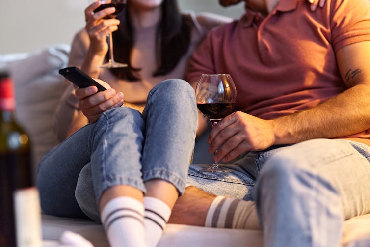 Bodies of man and woman sitting on a couch with tv remote and glasses of wine.