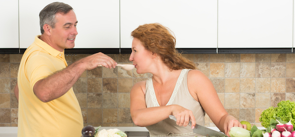 man giving his wife a taste of something from a spoon in a kitchen