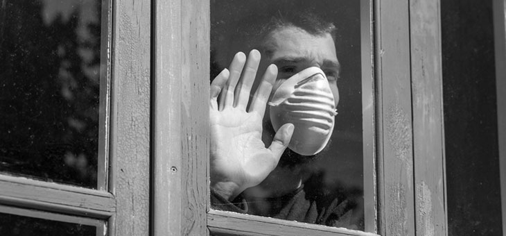 man wearing a mask in self isolation looking out the window