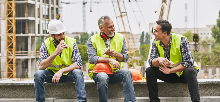 Group of builders in working uniform are eating sandwiches and talking while sitting on stone surface against construction site